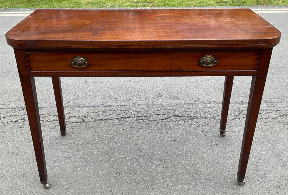 A Georgian mahogany card table featuring a rectangular fold-over top, a single long drawer with oval brass handles, tapered legs, and small brass castors.