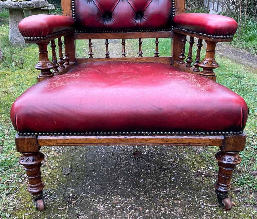 A Victorian Gothic Revival oak armchair featuring deep-buttoned red leather upholstery, carved finials, spindle arm supports, and turned legs with castors. View V