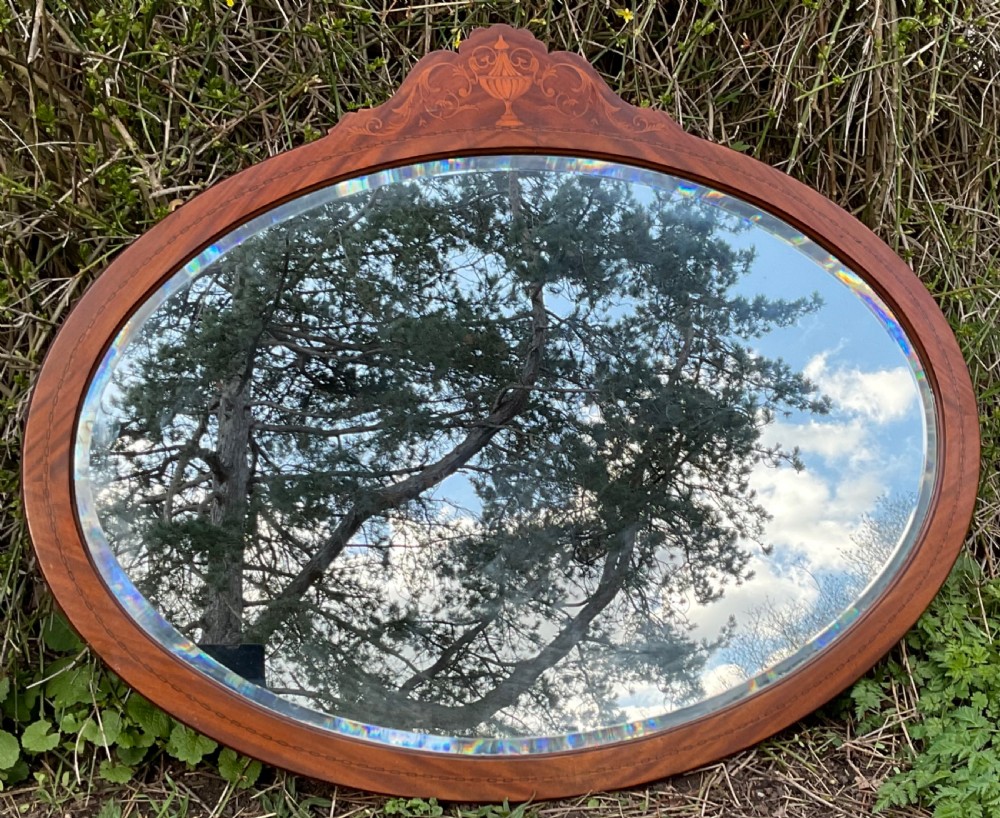 An oval mahogany framed wall mirror featuring a decorative pediment crest with marquetry urn inlay and bevelled glass.