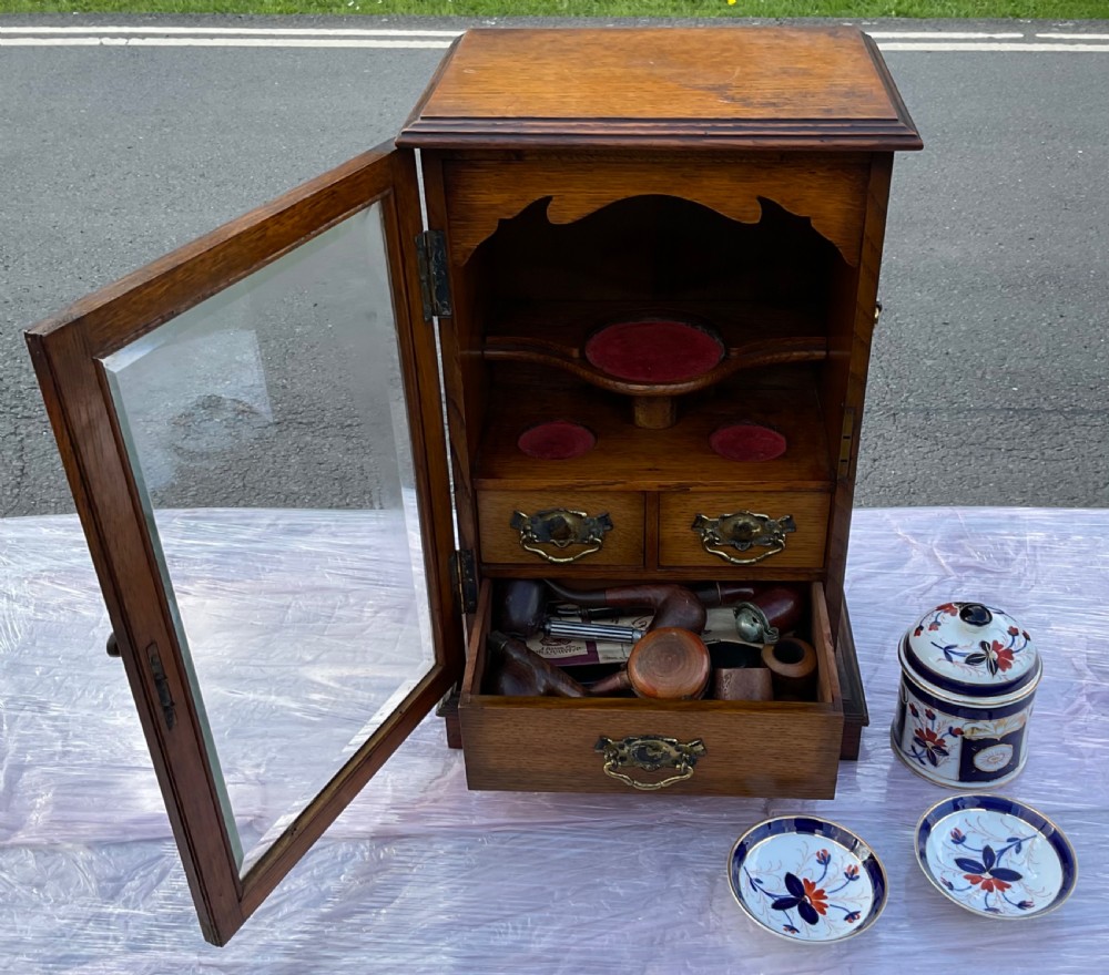 An Edwardian oak pipe smoking cabinet featuring a single glazed door revealing fitted shelves with a ceramic jar and dishes, above small internal drawers with brass handles. View X