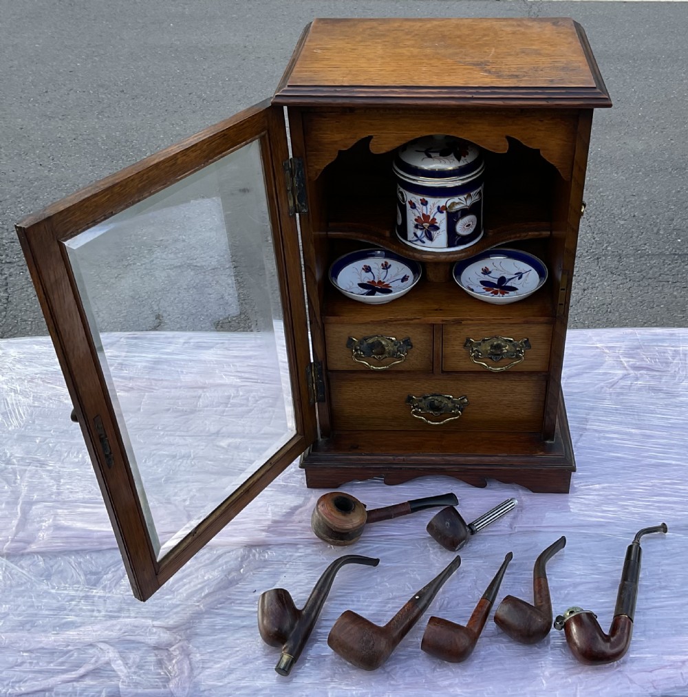 An Edwardian oak pipe smoking cabinet featuring a single glazed door revealing fitted shelves with a ceramic jar and dishes, above small internal drawers with brass handles. View XI