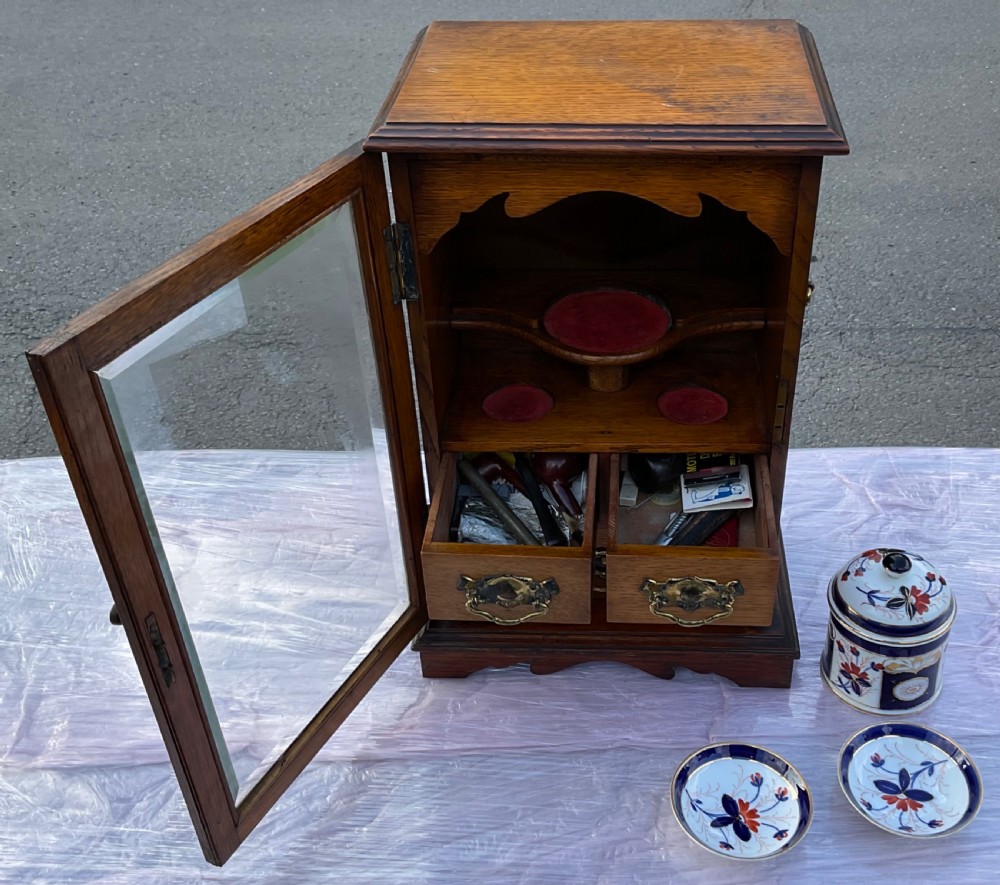 An Edwardian oak pipe smoking cabinet featuring a single glazed door revealing fitted shelves with a ceramic jar and dishes, above small internal drawers with brass handles. View XII