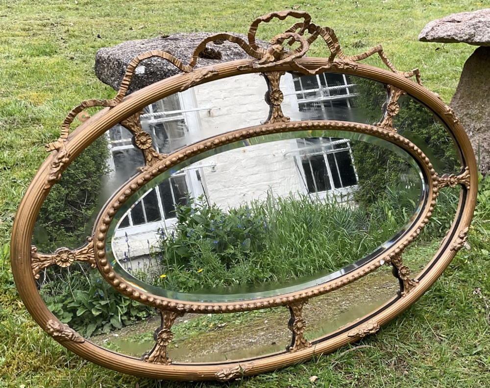 An horizontal oval giltwood wall mirror featuring a ribbon bow cresting, a beaded inner border, and floral rosette struts framing the bevelled glass. View IV