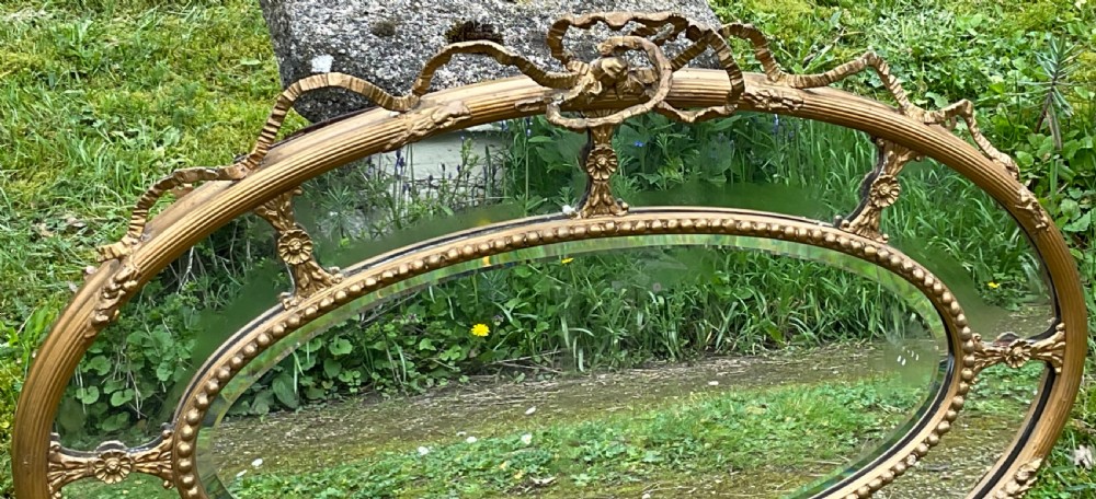 An horizontal oval giltwood wall mirror featuring a ribbon bow cresting, a beaded inner border, and floral rosette struts framing the bevelled glass. View VIII