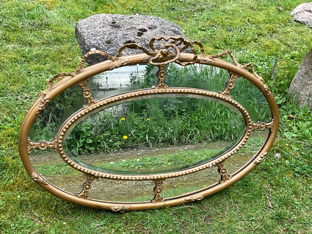 An horizontal oval giltwood wall mirror featuring a ribbon bow cresting, a beaded inner border, and floral rosette struts framing the bevelled glass. View IX