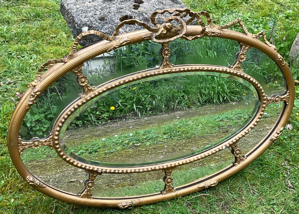 An horizontal oval giltwood wall mirror featuring a ribbon bow cresting, a beaded inner border, and floral rosette struts framing the bevelled glass. View X
