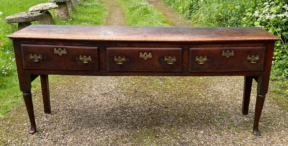 An 18th-century Georgian oak dresser base featuring three inline drawers with brass swan-neck handles, supported on slender turned legs with pad feet.