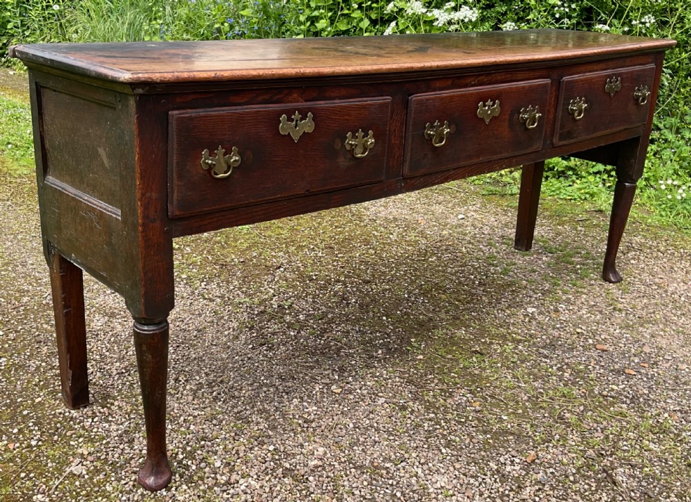 An 18th-century Georgian oak dresser base featuring three inline drawers with brass swan-neck handles, supported on slender turned legs with pad feet. View VI