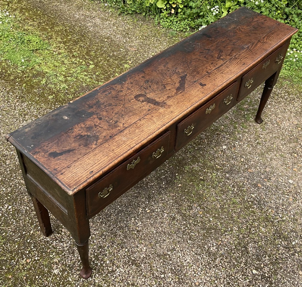 An 18th-century Georgian oak dresser base featuring three inline drawers with brass swan-neck handles, supported on slender turned legs with pad feet. View VII