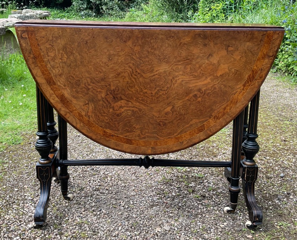 A Victorian drop-leaf table with a burr walnut oval top, ebonised turned legs, stretcher, and splayed feet on ceramic castors. View III