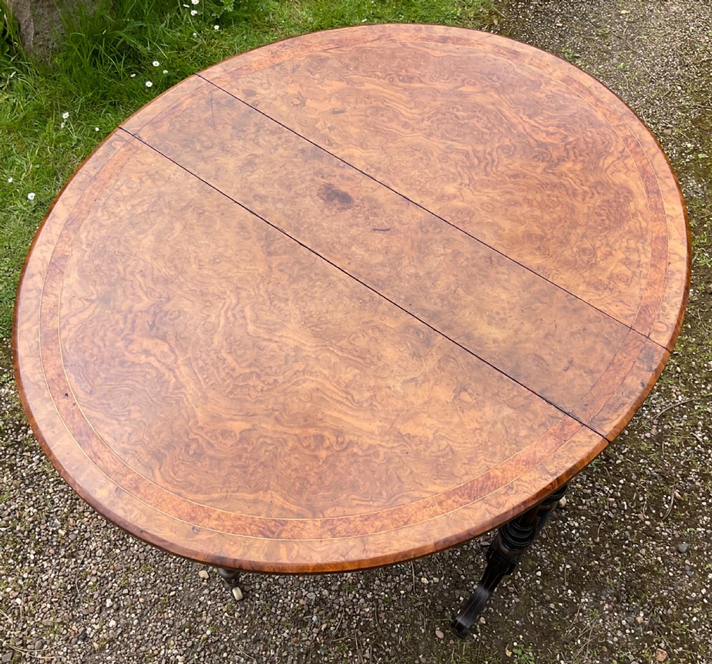 A Victorian drop-leaf table with a burr walnut oval top, ebonised turned legs, stretcher, and splayed feet on ceramic castors. View VI