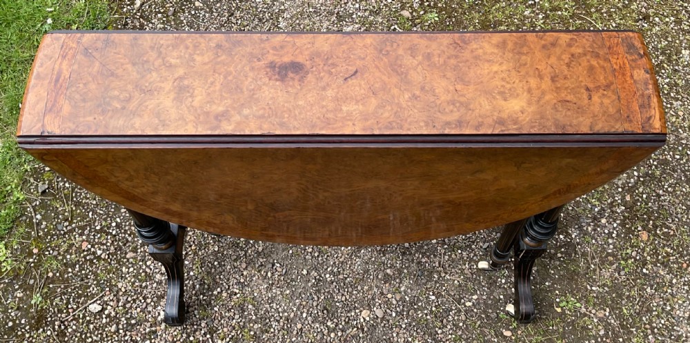 A Victorian drop-leaf table with a burr walnut oval top, ebonised turned legs, stretcher, and splayed feet on ceramic castors. View VII