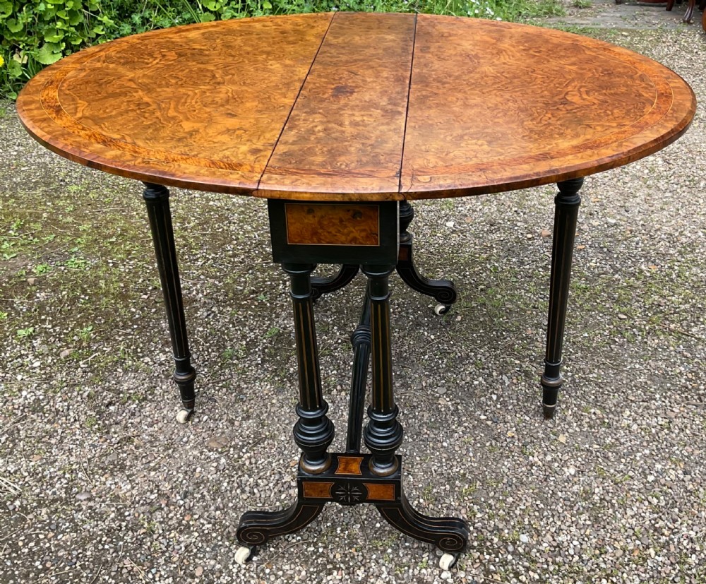 A Victorian drop-leaf table with a burr walnut oval top, ebonised turned legs, stretcher, and splayed feet on ceramic castors. View VIII