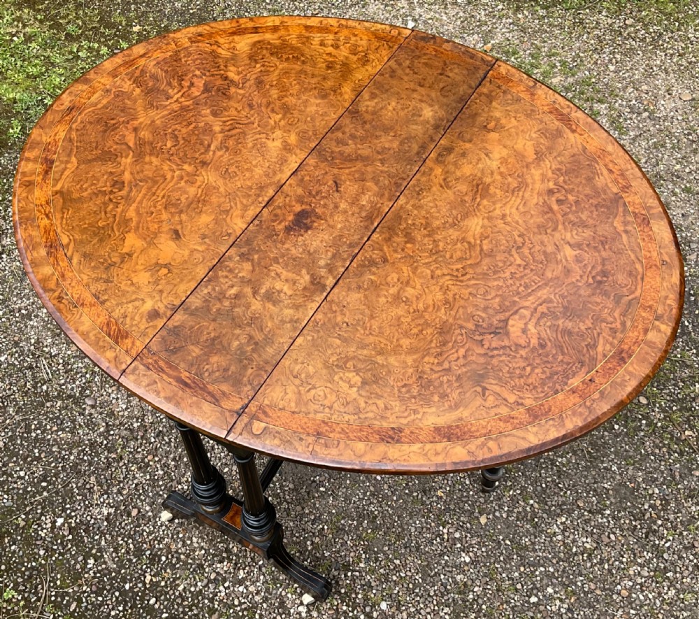 A Victorian drop-leaf table with a burr walnut oval top, ebonised turned legs, stretcher, and splayed feet on ceramic castors. View IX