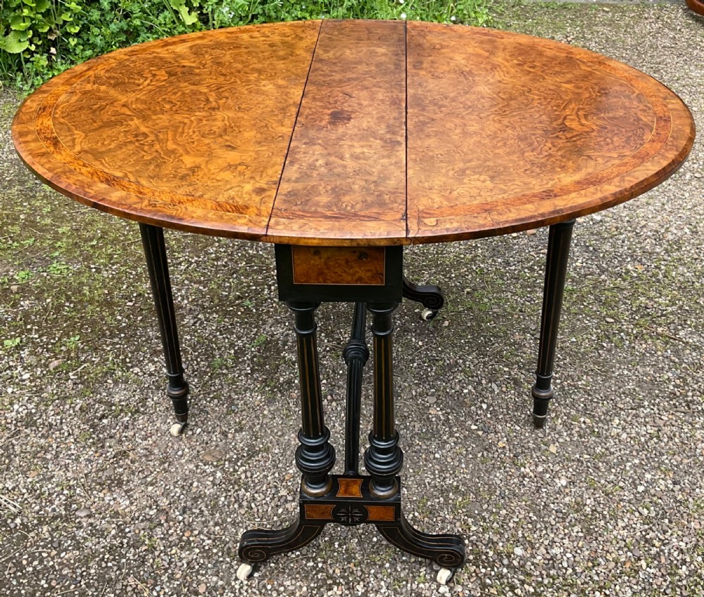 A Victorian drop-leaf table with a burr walnut oval top, ebonised turned legs, stretcher, and splayed feet on ceramic castors. View X