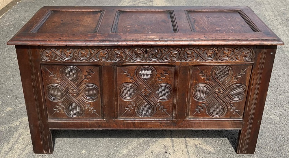 An antique dark oak blanket chest featuring three relief-carved front panels, a carved upper frieze, and a three-panel hinged top. View II
