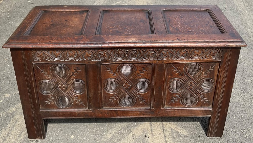An antique dark oak blanket chest featuring three relief-carved front panels, a carved upper frieze, and a three-panel hinged top. View III