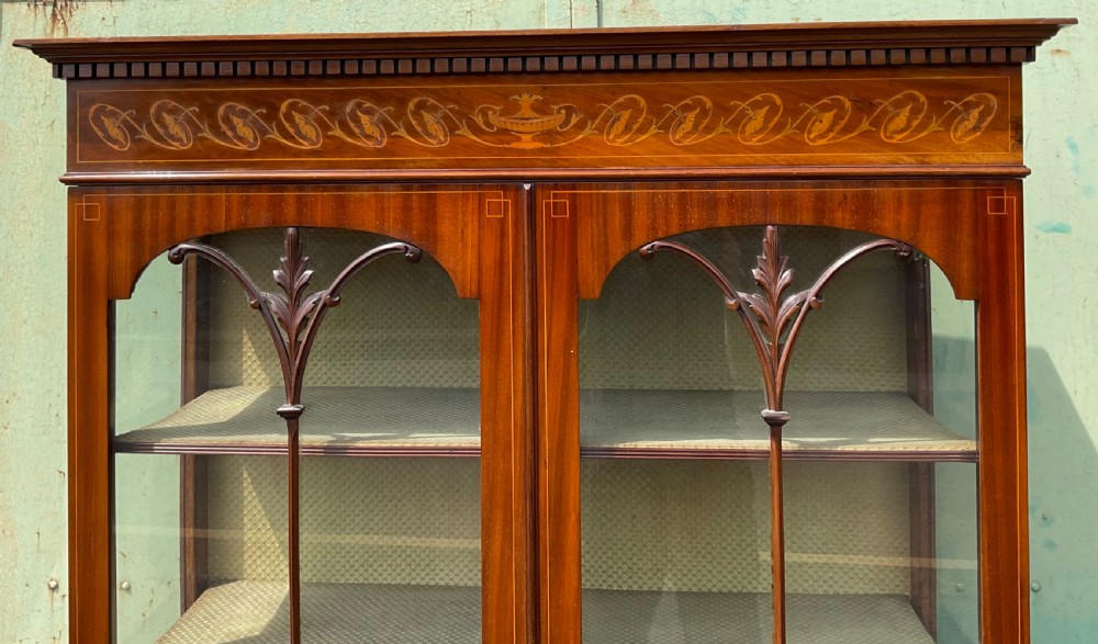 An Edwardian inlaid mahogany display cabinet featuring two glazed doors with decorative glazing bars, an inlaid urn frieze, and square tapered legs. View V