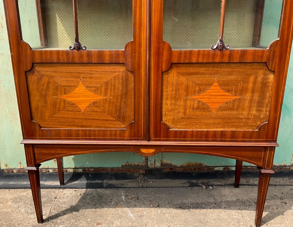 An Edwardian inlaid mahogany display cabinet featuring two glazed doors with decorative glazing bars, an inlaid urn frieze, and square tapered legs. View VI