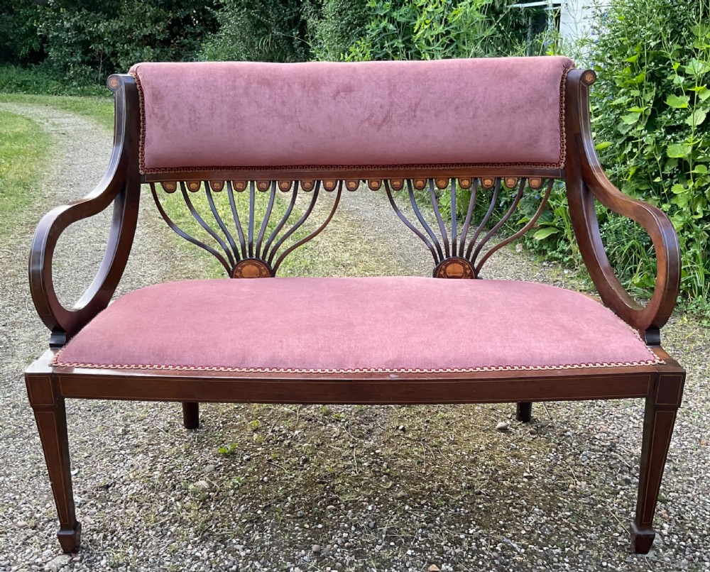 An Edwardian inlaid mahogany salon settee featuring a padded dusty pink velvet backrest and seat, flanked by sweeping arms and supported by splats with fan-shaped inlay.