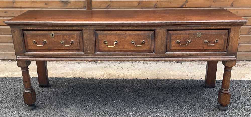 An antique oak dresser base featuring three panelled drawers with brass swan-neck handles and turned front legs.