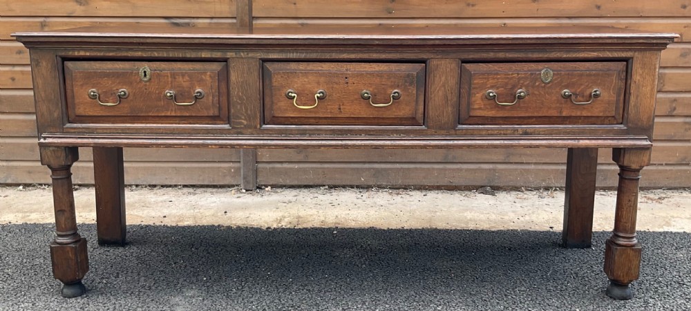 An antique oak dresser base featuring three panelled drawers with brass swan-neck handles and turned front legs. View II