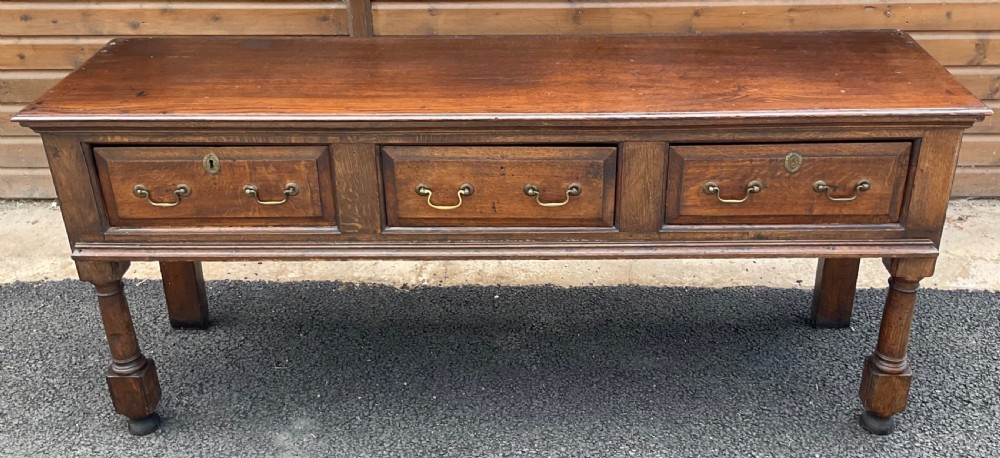 An antique oak dresser base featuring three panelled drawers with brass swan-neck handles and turned front legs. View III