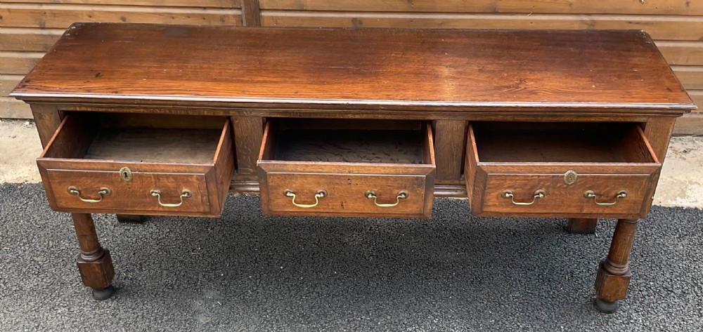 An antique oak dresser base featuring three panelled drawers with brass swan-neck handles and turned front legs. View IV