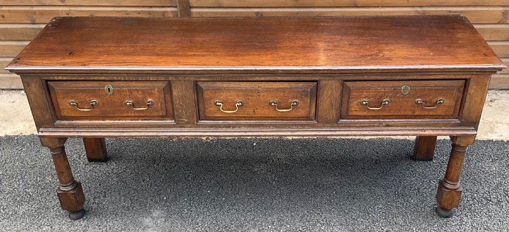 An antique oak dresser base featuring three panelled drawers with brass swan-neck handles and turned front legs. View V