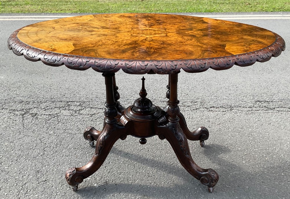 An oval Victorian centre table crafted from figure-grained burr walnut with floral marquetry inlay, carved scalloped edges, and four carved pedestal legs on castors.