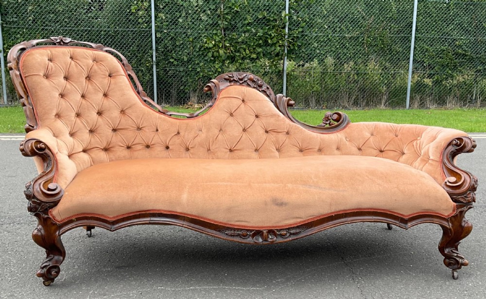An antique Victorian mahogany chaise longue featuring carved show-wood scrollwork, an asymmetrical backrest, and button-tufted peach velvet upholstery.