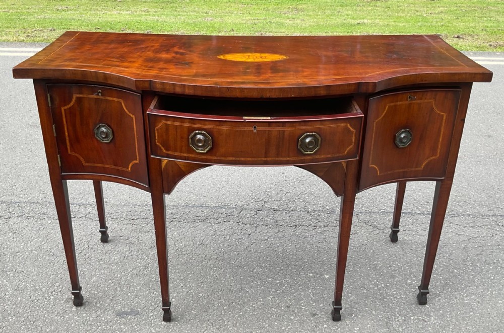 A Georgian mahogany bowfront sideboard featuring marquetry inlays, a central drawer, flanking cupboard doors, and tapered legs. View VII
