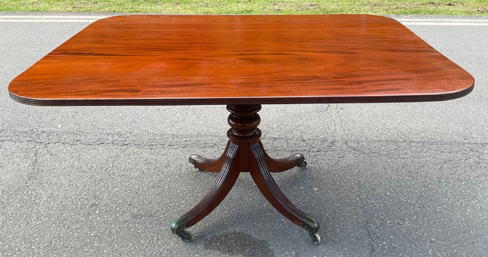 A Georgian mahogany pedestal table featuring a rectangular top with rounded edges, a turned central column, and four splayed reeded legs on brass castors.