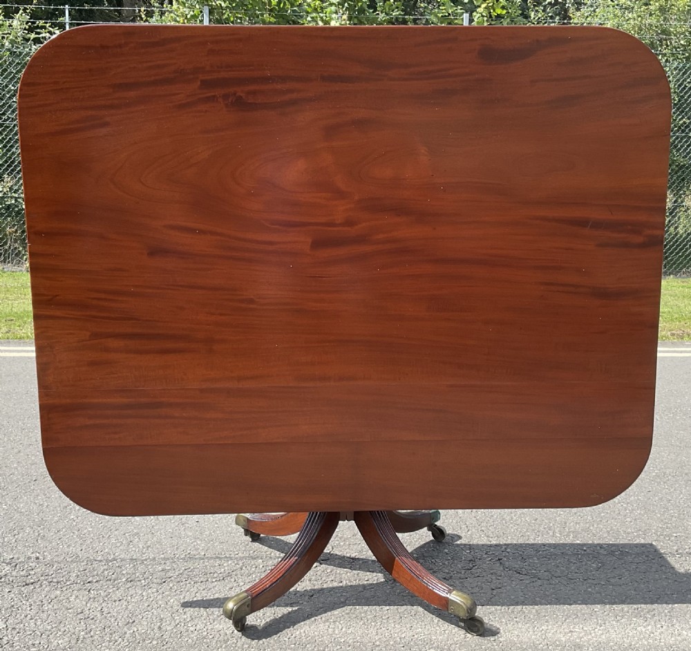 A Georgian mahogany pedestal table featuring a rectangular top with rounded edges, a turned central column, and four splayed reeded legs on brass castors. View II