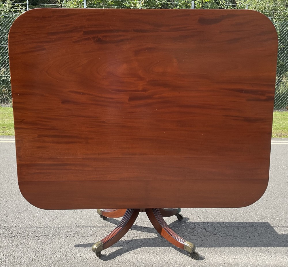 A Georgian mahogany pedestal table featuring a rectangular top with rounded edges, a turned central column, and four splayed reeded legs on brass castors. View IV