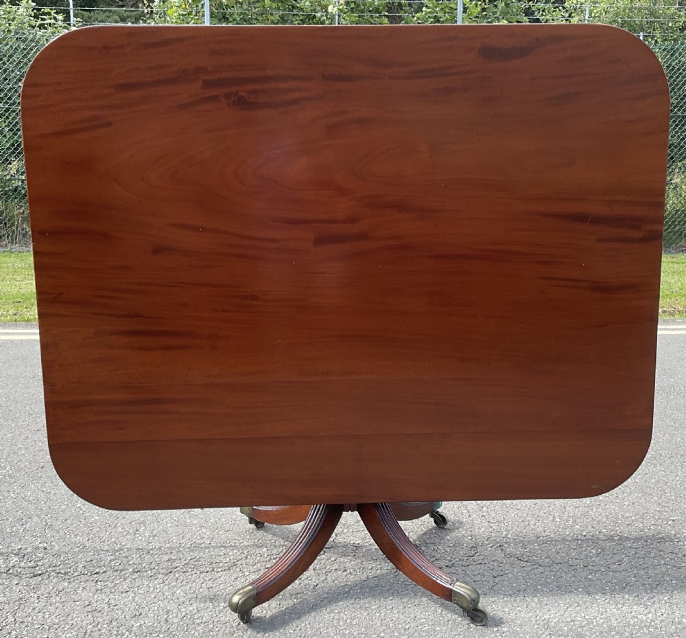 A Georgian mahogany pedestal table featuring a rectangular top with rounded edges, a turned central column, and four splayed reeded legs on brass castors. View V