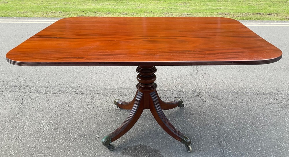 A Georgian mahogany pedestal table featuring a rectangular top with rounded edges, a turned central column, and four splayed reeded legs on brass castors. View VIII