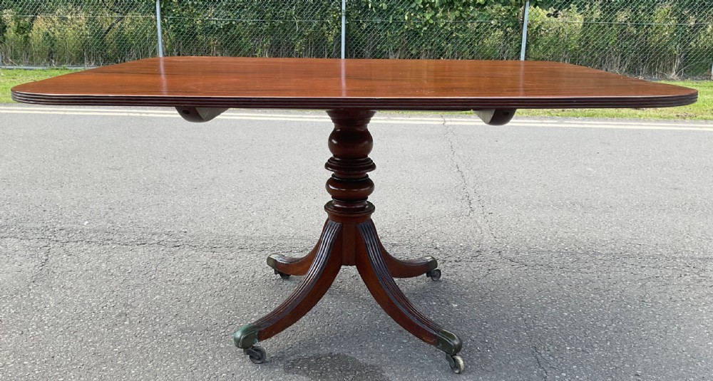 A Georgian mahogany pedestal table featuring a rectangular top with rounded edges, a turned central column, and four splayed reeded legs on brass castors. View X