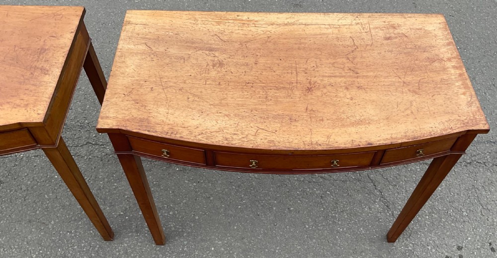 A pair of Georgian-style mahogany side tables featuring gentle bowfronts, drawer fronts with brass ring handles, and square tapered legs. View X