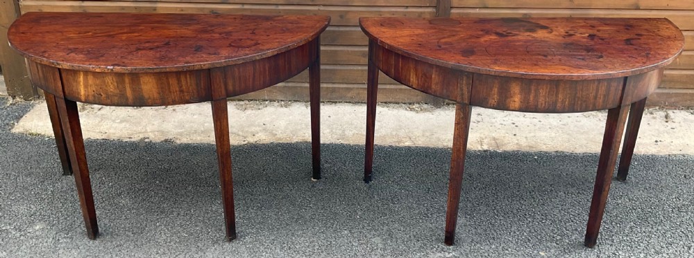 A pair of Georgian dark mahogany D-end console tables with semi-circular tops, veneered aprons, and straight tapered legs.