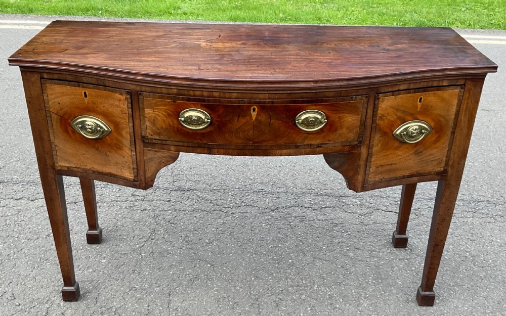 A Georgian mahogany bowfront sideboard featuring flame veneer drawer fronts, brass oval handles, and tapered square legs.