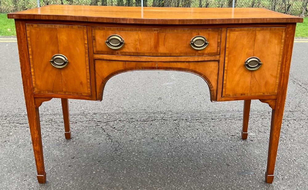 A Georgian satinwood bowfront sideboard featuring a central drawer above a shallow arch, flanked by two side cupboards with brass oval drop handles and supported on tapered square legs.