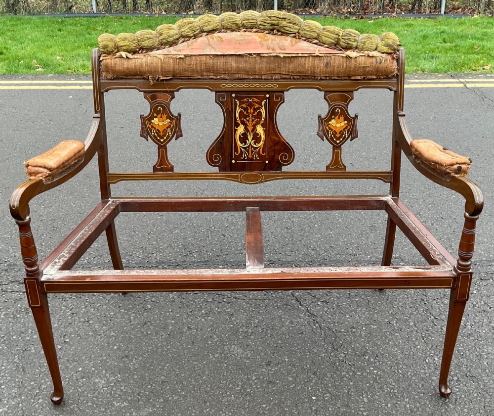 An antique Edwardian mahogany salon settee frame featuring intricate marquetry inlays on the backrest splats, padded armrests, and slender tapered legs.