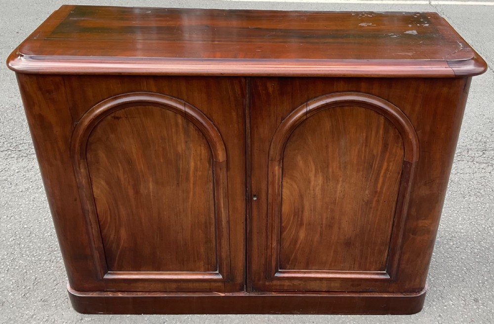 A Victorian dark warm brown mahogany side cupboard featuring two arched panel doors and a solid plinth base.
