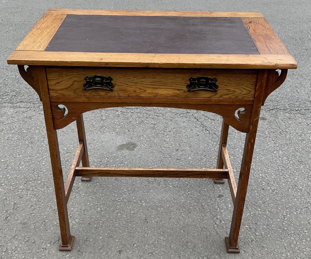 An Art Nouveau solid oak writing table featuring a dark brown leather inset top, pierced corner brackets, and a single drawer with decorative metal handles.