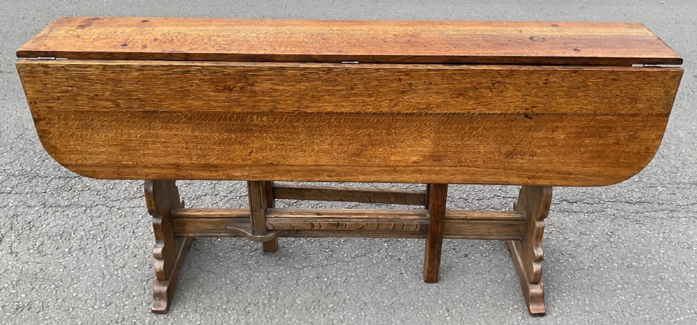 A long, narrow antique Arts and Crafts oak gateleg dining table with drop-leaf sides and shaped trestle end supports.