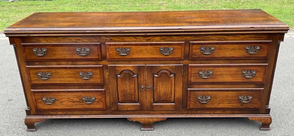 A low Georgian-style oak sideboard featuring multiple crossbanded drawers surrounding a central two-door cupboard with arched fielded panels and brass swan-neck handles.