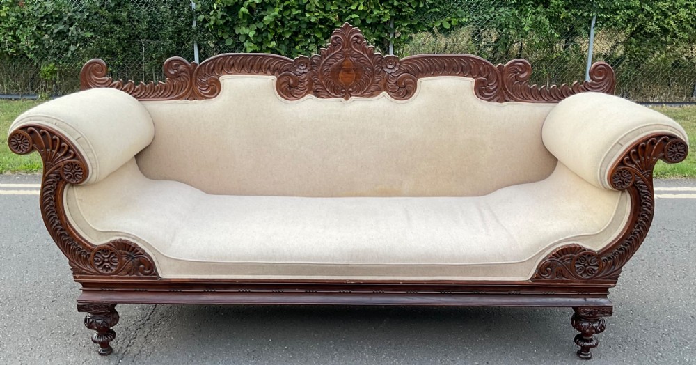 An antique Regency carved mahogany four-seater settee featuring rolled scroll arms, floral cresting, and cream upholstery.