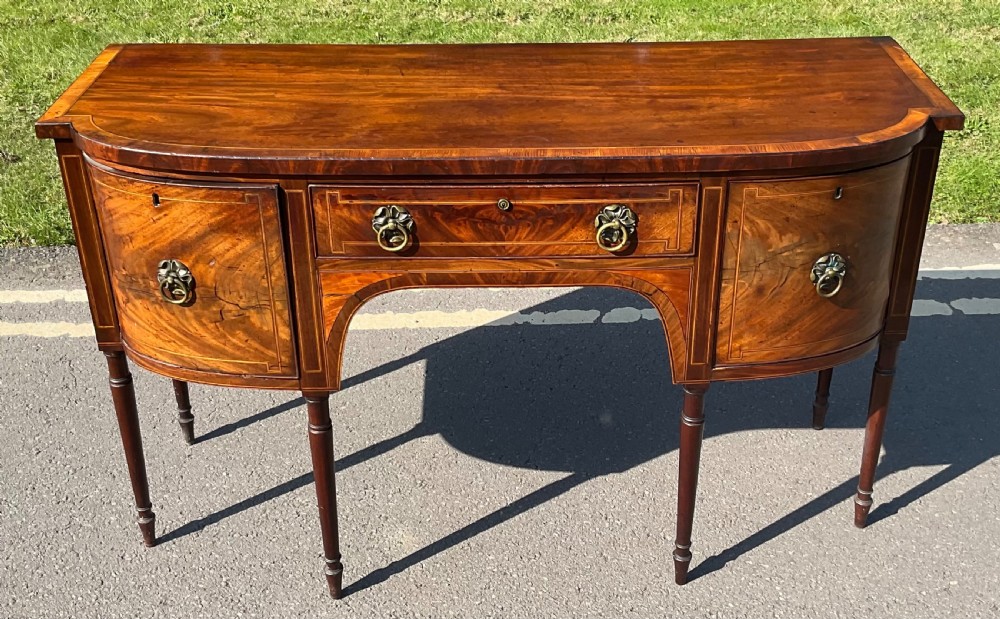 A Georgian mahogany bowfront sideboard featuring boxwood stringing inlay, brass ring handles, and turned tapered legs.