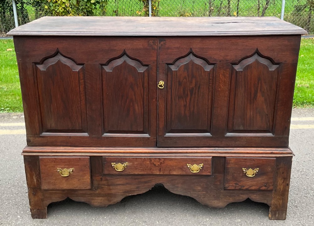 A Georgian dark oak cupboard featuring ogee arched fielded panels on two upper doors and brass handle hardware on lower drawers.
